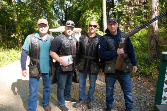 Four men in shooting vests with a shotgun