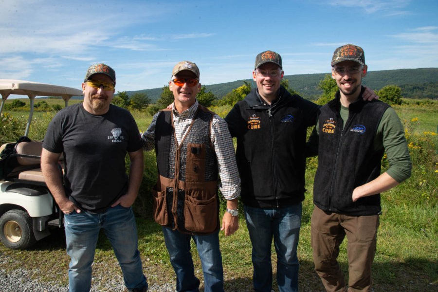Four men smiling outdoors in caps and vests