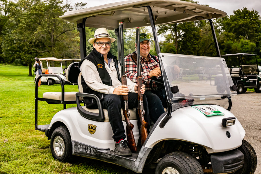 Two men seated in golf cart holding shotguns