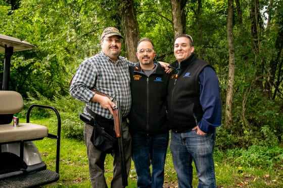 Three men standing in woods, one with shotgun