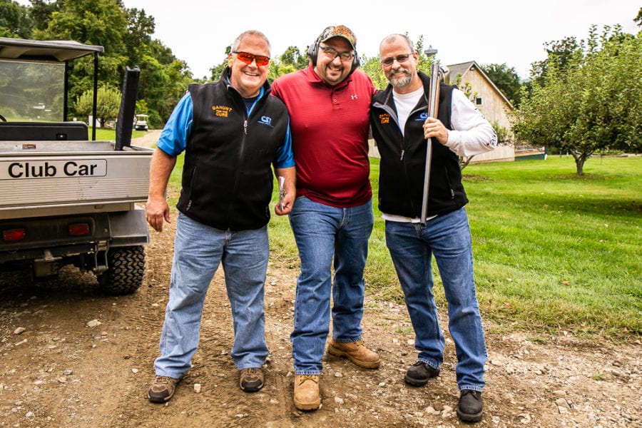 Three men posing outdoors, one holding shotgun