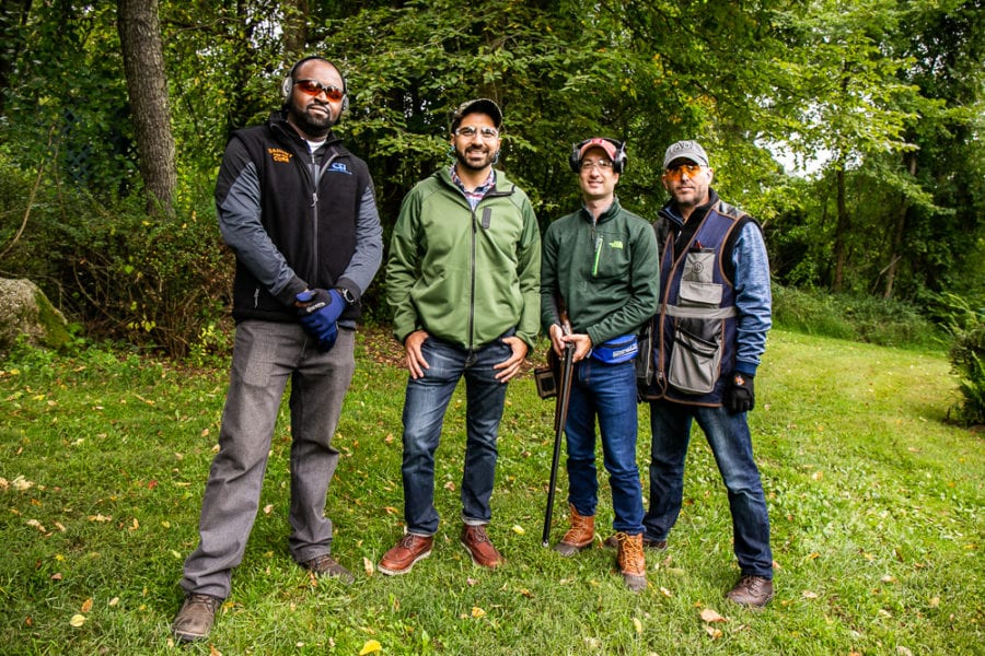Four men outdoors, one holding a shotgun