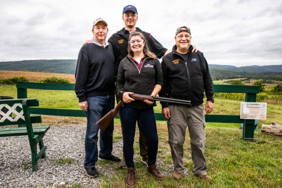 Four people posing outdoors, woman holding shotgun