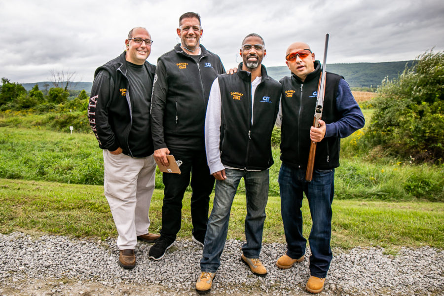 Four men posing outdoors, one holding shotgun