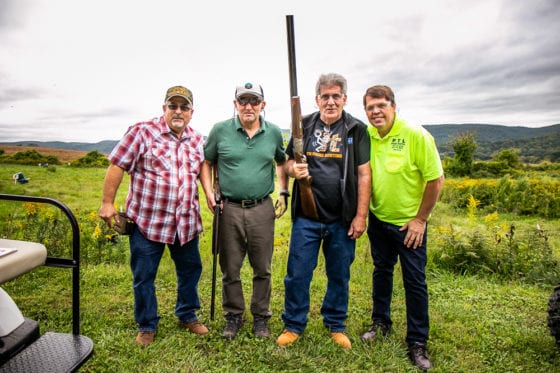 Four men outdoors, one holding a shotgun