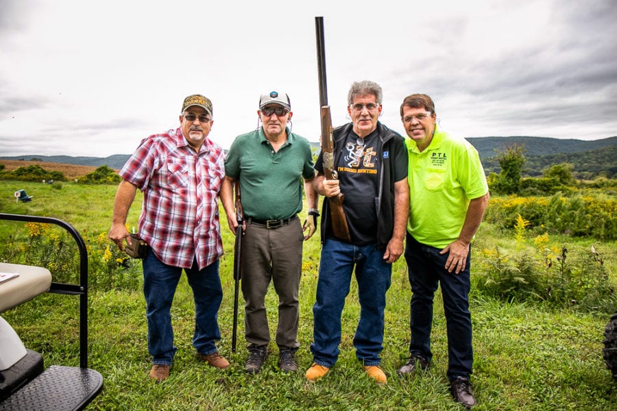 Four men outdoors, one holding a shotgun