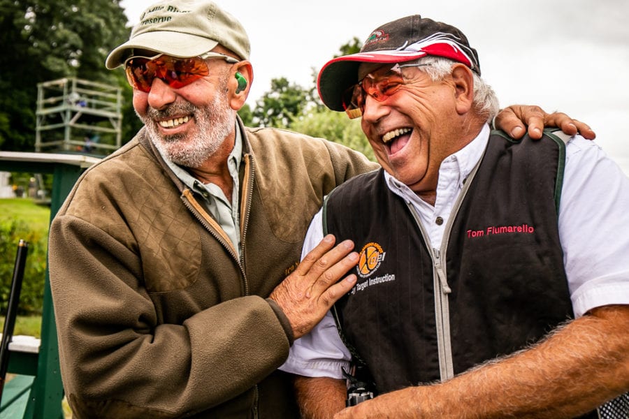 Two men laughing outdoors wearing sunglasses, caps