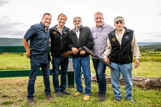Five men outdoors smiling, one holding shotgun